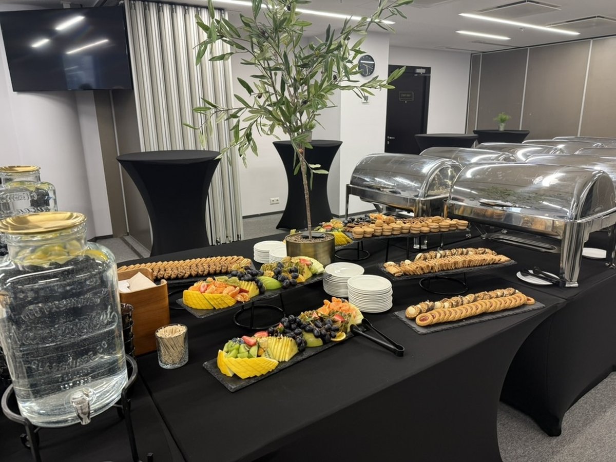 Konverentsi musta linaga toidulaud. Alused puuviljadega, väikesed küpsised, soolased suupisted ja metallist kaanega kausid. Taustal mustade katetega kõrged seisulauad ja veekannud.