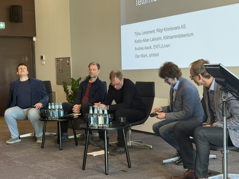 Viis meest istuvad mustades tugitoolides paneelarutelu ajal. Nende vahel väikesed ümmargused lauad veepudelitega. Taustal ekraanil panelistide nimed: Tõnu Lensment (Riigi Kinnisvara AS), Kaido-Allan Lainurm (Kliimaministeerium), Andres Aavik (EKFL/Liven) ja Ülar Mark (arhitekt). Vasakul seisab must kõlar.