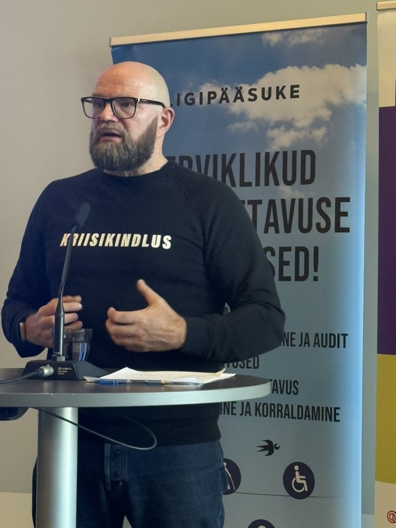 Priit Laos Kriisikindlus OÜ-st kõneleb ümarlaua taga, käed žestikuleerimas. Tema ees laual mikrofon ja paberid. Taustal Ligipääsuke reklaamstend teenuste nimekirjaga.